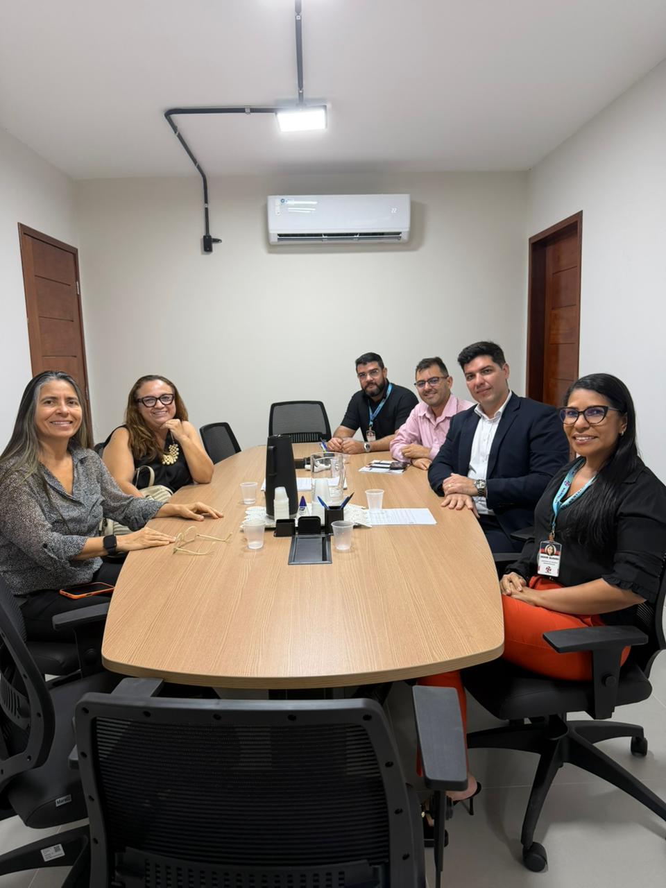 Seis pessoas, sendo três mulheres e três homens, estão sentadas sorrindo ao redor de uma grande mesa de reunião de madeira em um escritório com paredes brancas.