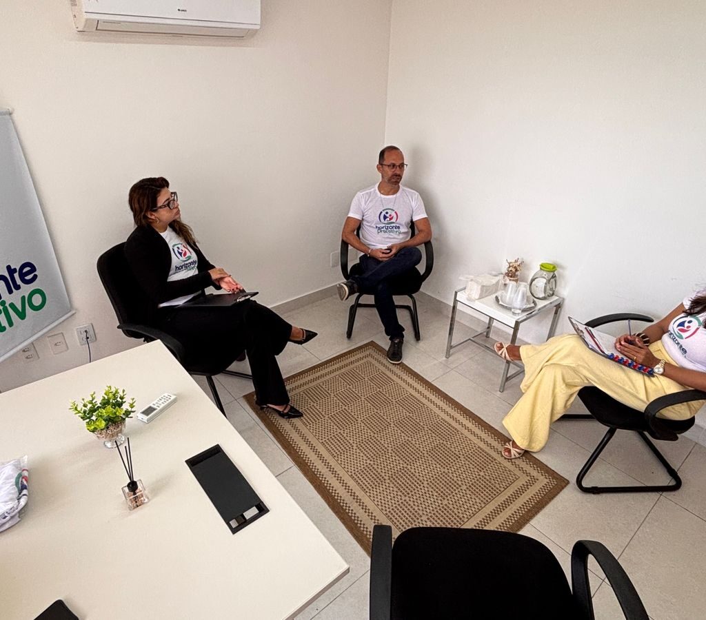 Três pessoas participam de uma reunião em uma sala de escritório com paredes brancas, onde se destaca um banner com a logo "horizonte protetivo".