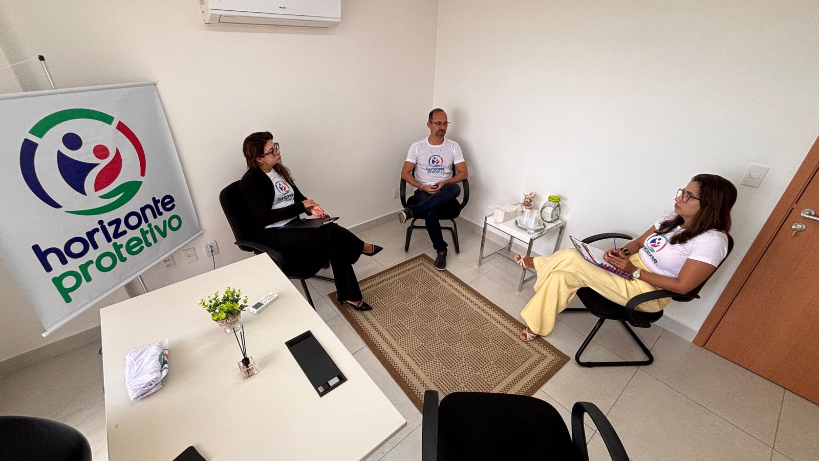 Três pessoas participam de uma reunião em uma sala de escritório com paredes brancas, onde se destaca um banner com a logo "horizonte protetivo".