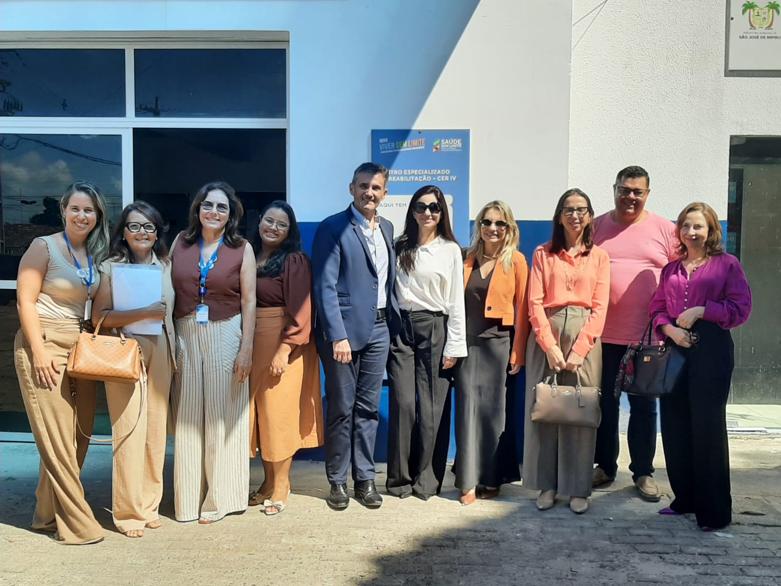 grupo de dez pessoas, vestidas com trajes casuais e formais, sorrindo e posadas lado a lado em frente à entrada de um prédio público