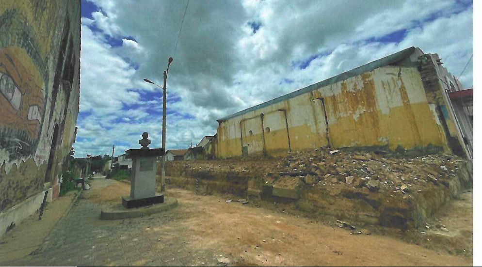 Fotografia em ângulo aberto de uma rua de terra com um busto em um pedestal de concreto, ladeada por edifícios antigos com paredes desgastadas e pichadas, sob um céu nublado.