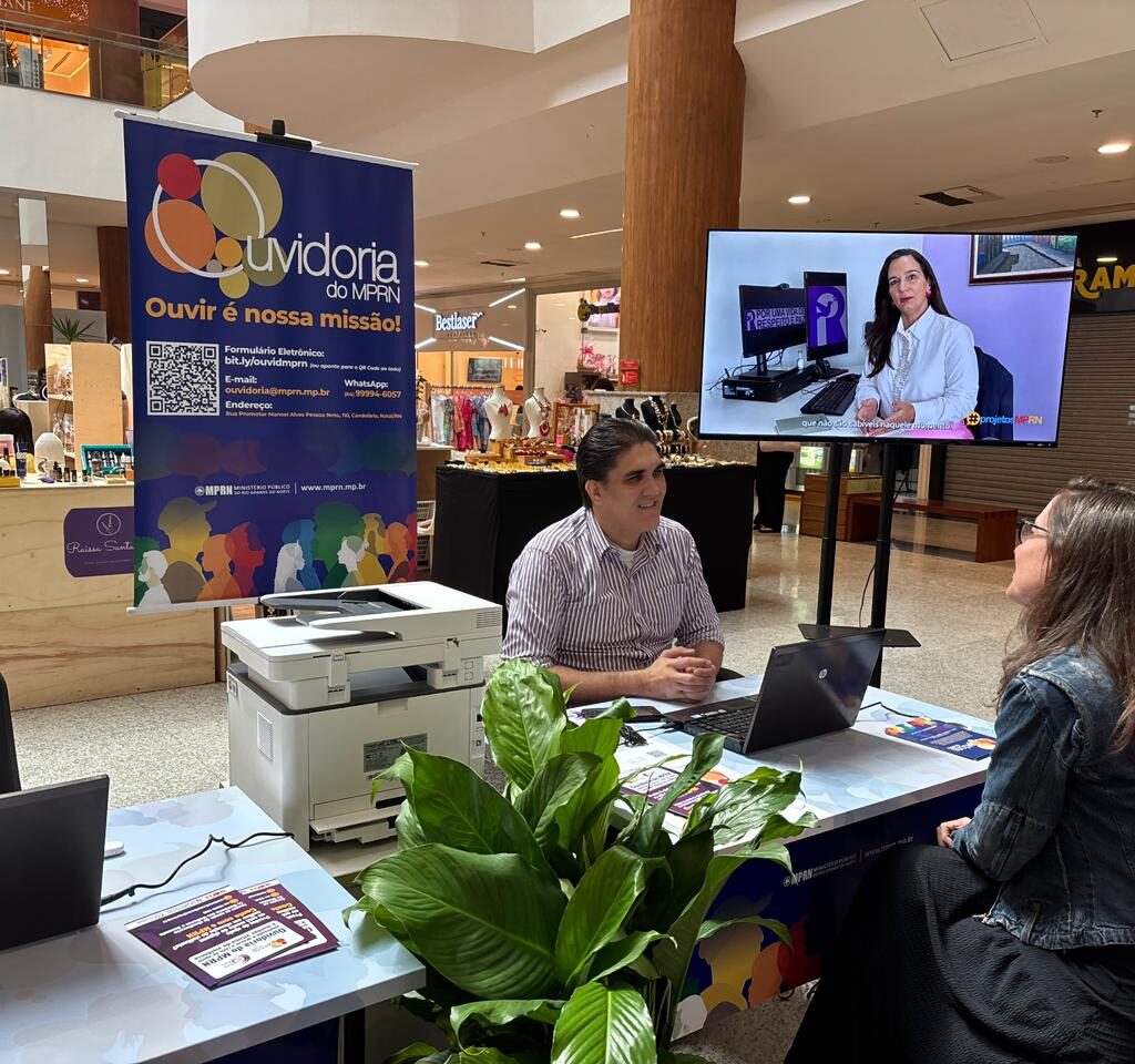Homem e mulher conversam sentados em um estande de atendimento da Ouvidoria do MPRN, estruturado com computadores, impressora e um painel informativo, no corredor de um shopping.