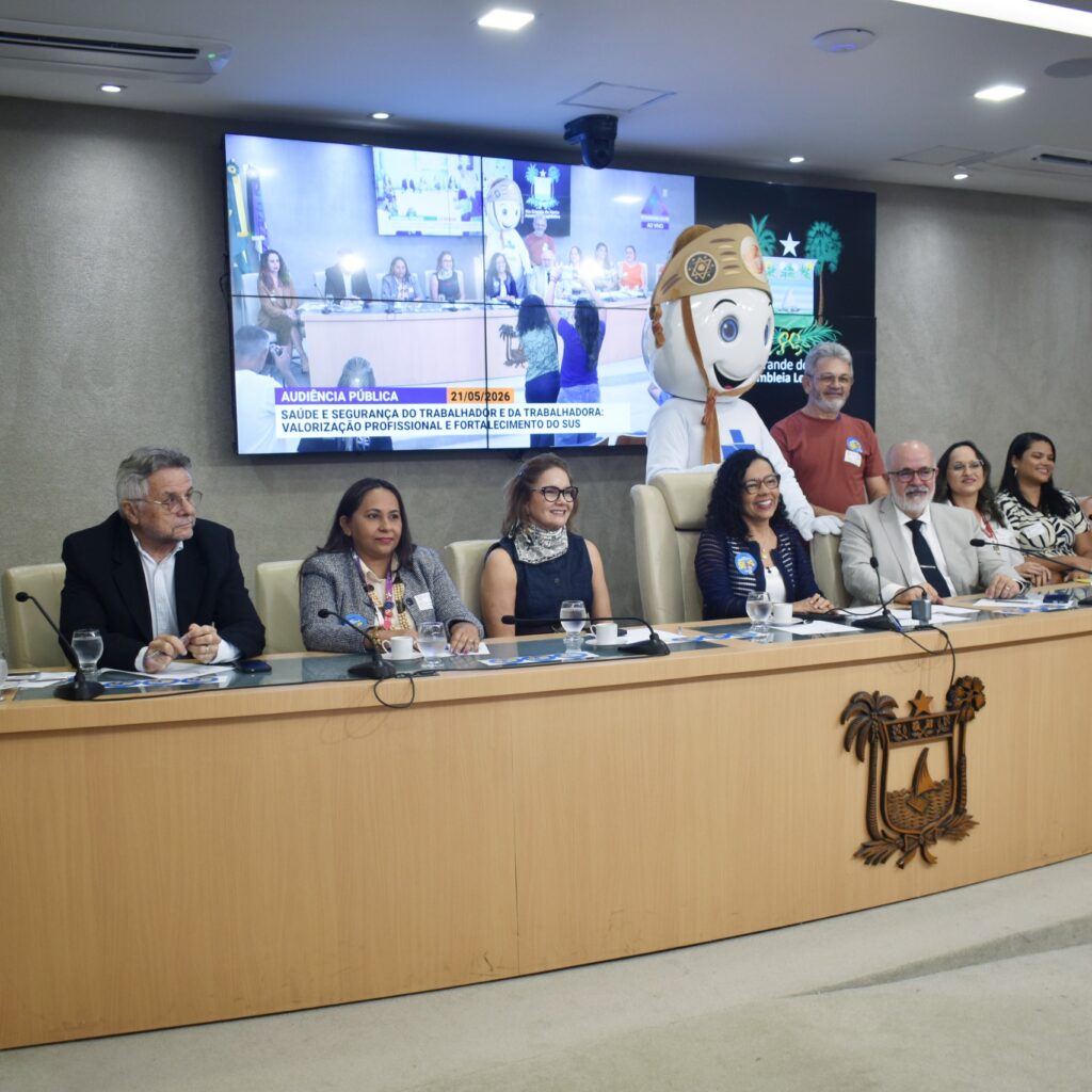 audiência pública em um auditório, mostrando uma mesa com diversas autoridades sentadas e um boneco mascote em pé ao fundo, posicionados em frente a um grande painel que exibe a transmissão do evento.