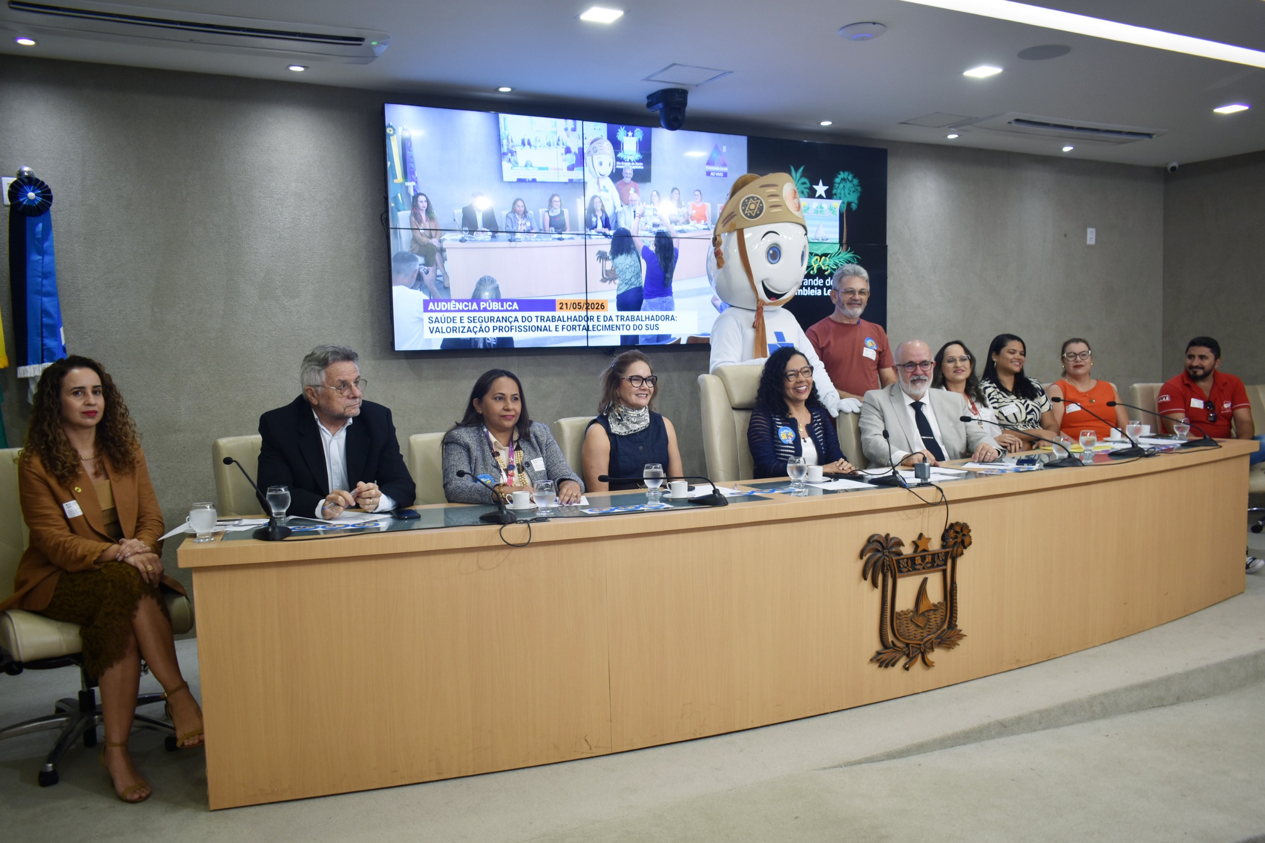 audiência pública em um auditório, mostrando uma mesa com diversas autoridades sentadas e um boneco mascote em pé ao fundo, posicionados em frente a um grande painel que exibe a transmissão do evento.