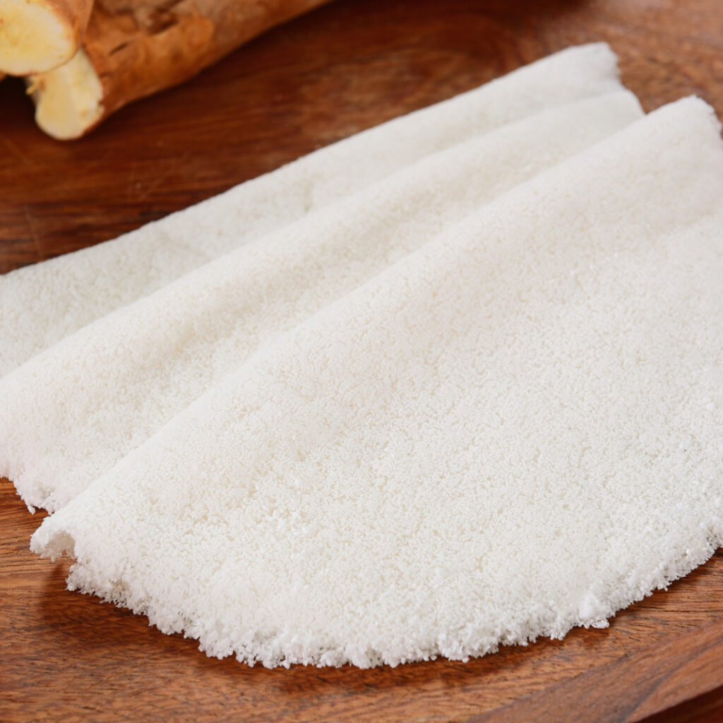 Uma tapioca branca, dobrada ao meio em formato de meia-lua, está disposta sobre uma tábua de madeira escura com pedaços de mandioca ao fundo.