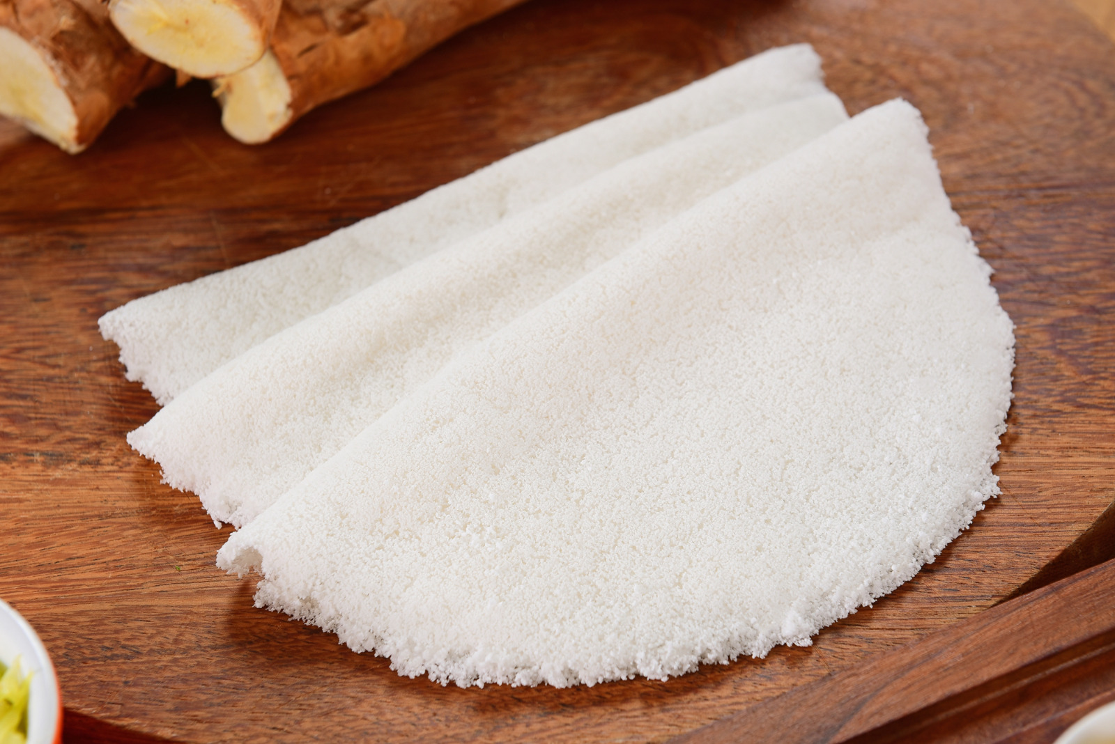 Uma tapioca branca, dobrada ao meio em formato de meia-lua, está disposta sobre uma tábua de madeira escura com pedaços de mandioca ao fundo.