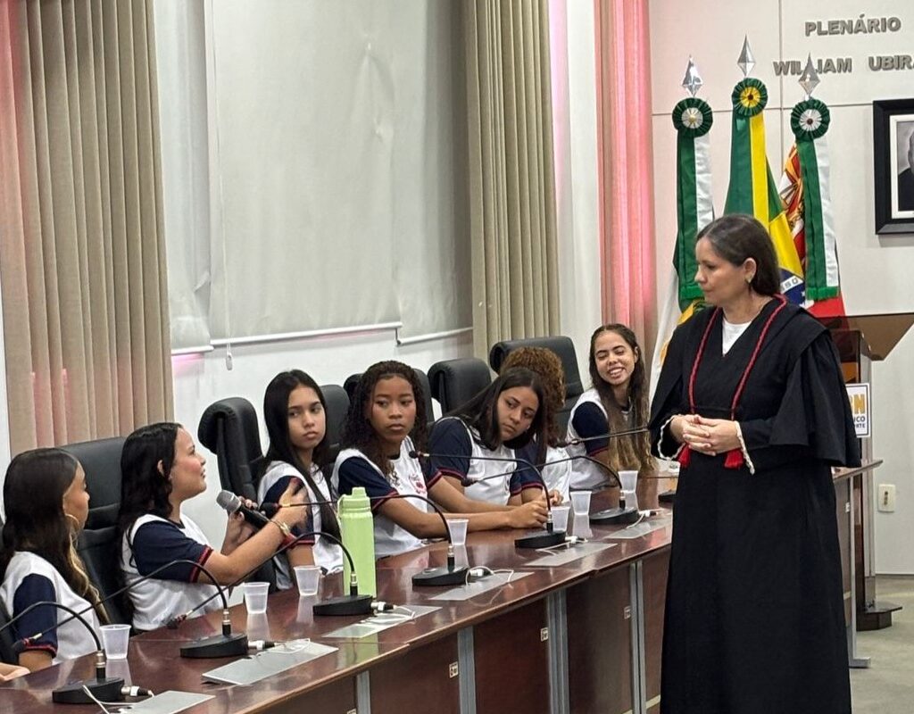 Uma mulher vestindo toga preta conversa com um grupo de estudantes uniformizados sentados ao redor de uma longa mesa de plenário.