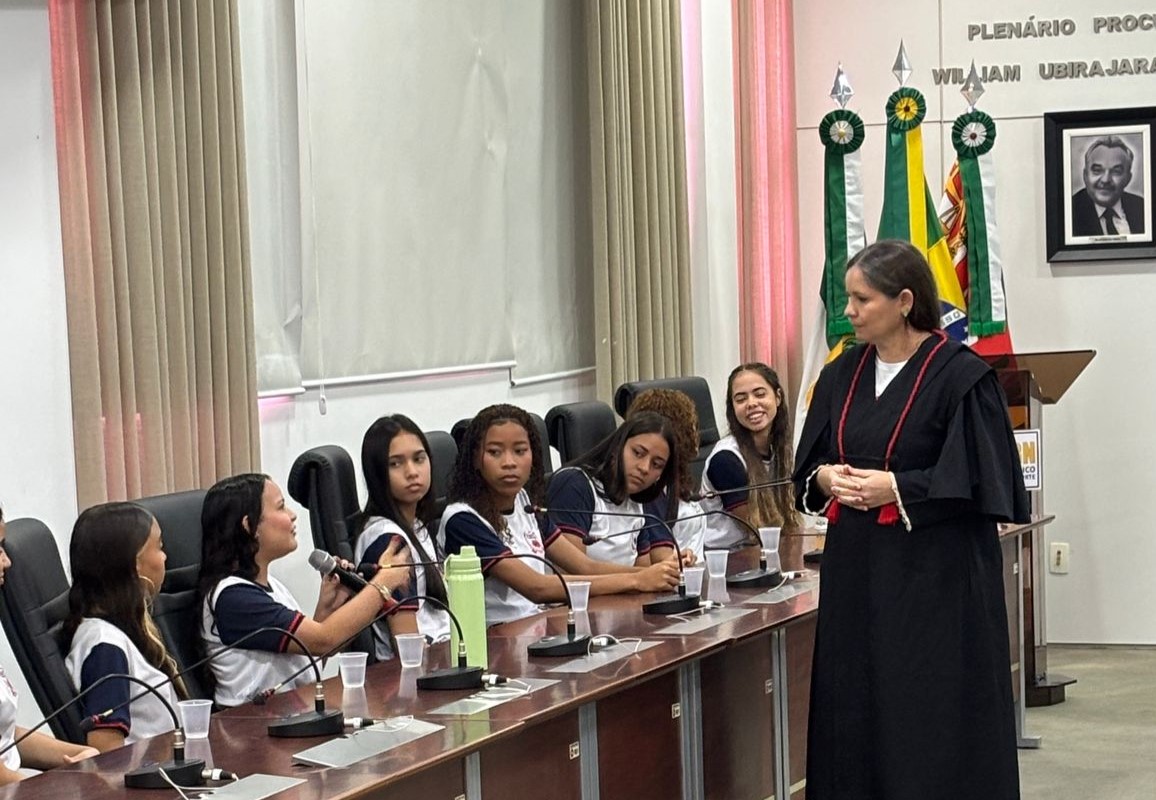 Uma mulher vestindo toga preta conversa com um grupo de estudantes uniformizados sentados ao redor de uma longa mesa de plenário.
