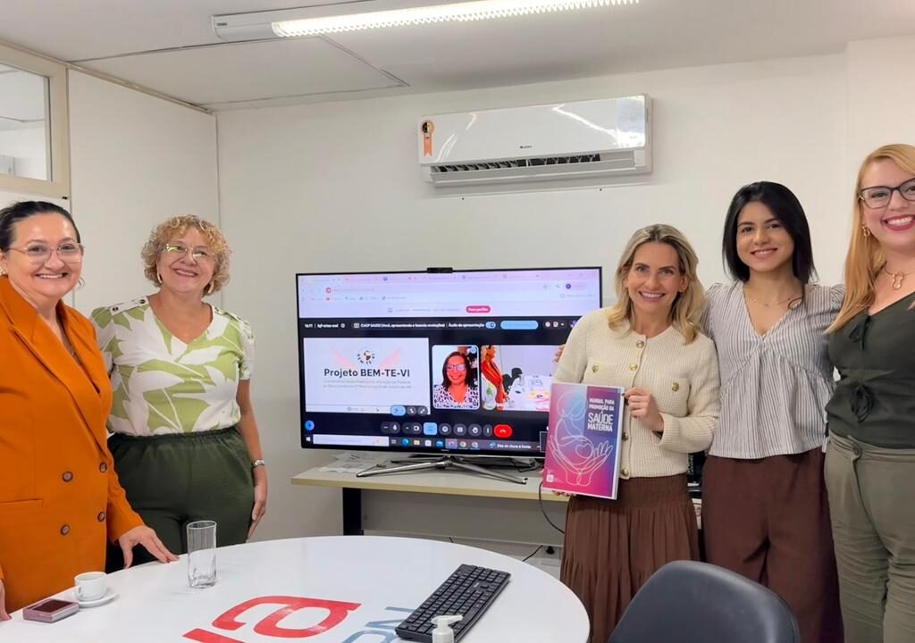 Cinco mulheres sorriem em uma sala de reuniões ao redor de uma TV que exibe uma videoconferência do "Projeto BEM-TE-VI", enquanto uma delas segura uma cartilha sobre saúde materna.