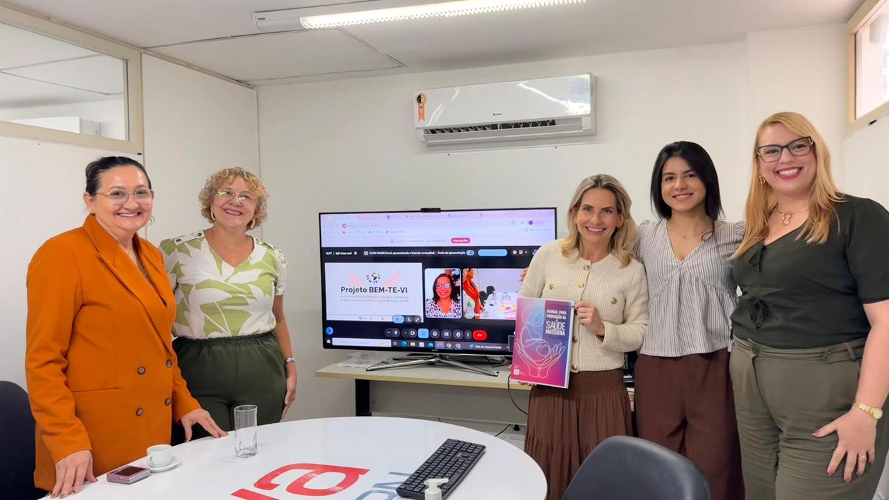 Cinco mulheres sorriem em uma sala de reuniões ao redor de uma TV que exibe uma videoconferência do "Projeto BEM-TE-VI", enquanto uma delas segura uma cartilha sobre saúde materna.