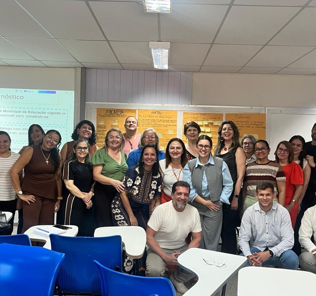 Grupo de cerca de vinte pessoas posa sorridente para foto em uma sala de aula com carteiras escolares azuis em primeiro plano e uma tela de projeção ao fundo.