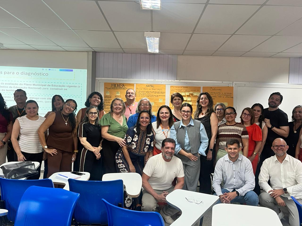 Grupo de cerca de vinte pessoas posa sorridente para foto em uma sala de aula com carteiras escolares azuis em primeiro plano e uma tela de projeção ao fundo.
