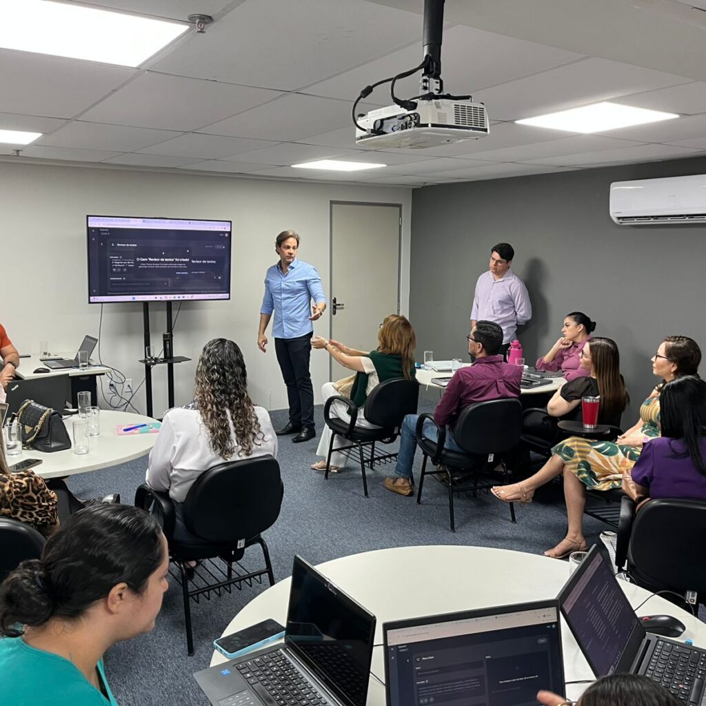 Um homem de camisa azul clara em pé apresenta uma palestra sobre inteligência artificial para um grupo de pessoas sentadas em mesas redondas em uma sala de reuniões.
