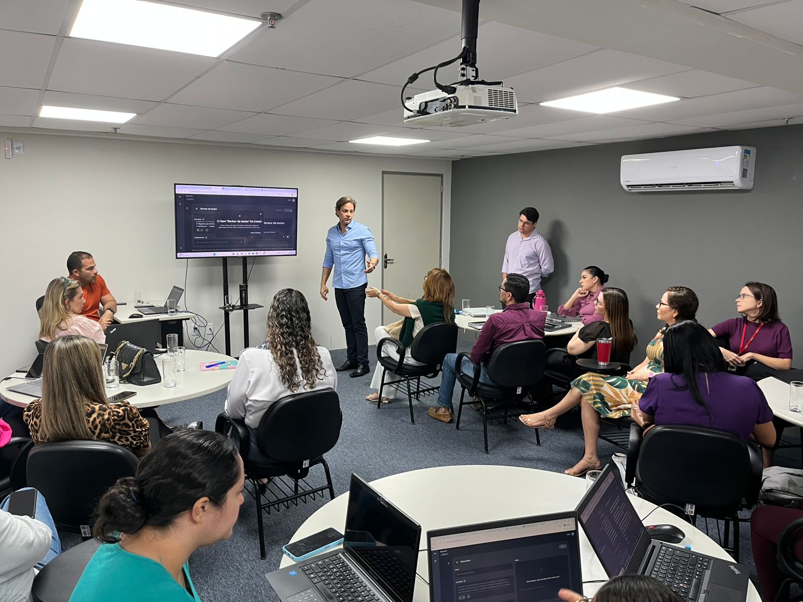 Um homem de camisa azul clara em pé apresenta uma palestra sobre inteligência artificial para um grupo de pessoas sentadas em mesas redondas em uma sala de reuniões.