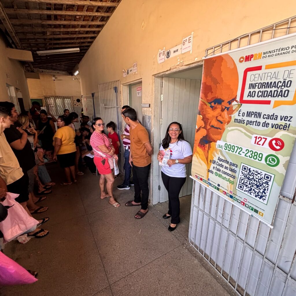 corredor de escola com pessoas e servidora do MP ao lado de banner indincando atendimento