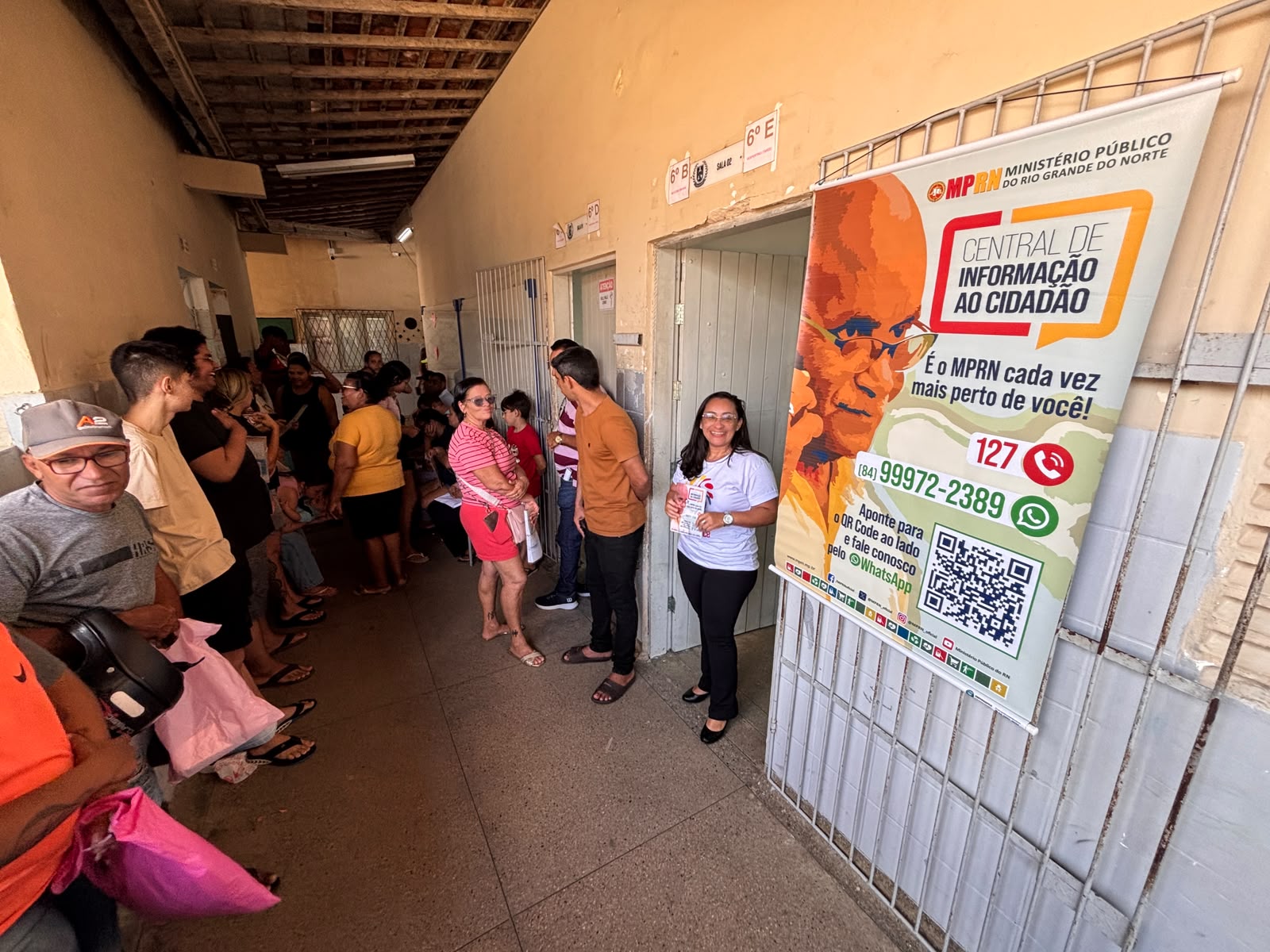 corredor de escola com pessoas e servidora do MP ao lado de banner indincando atendimento