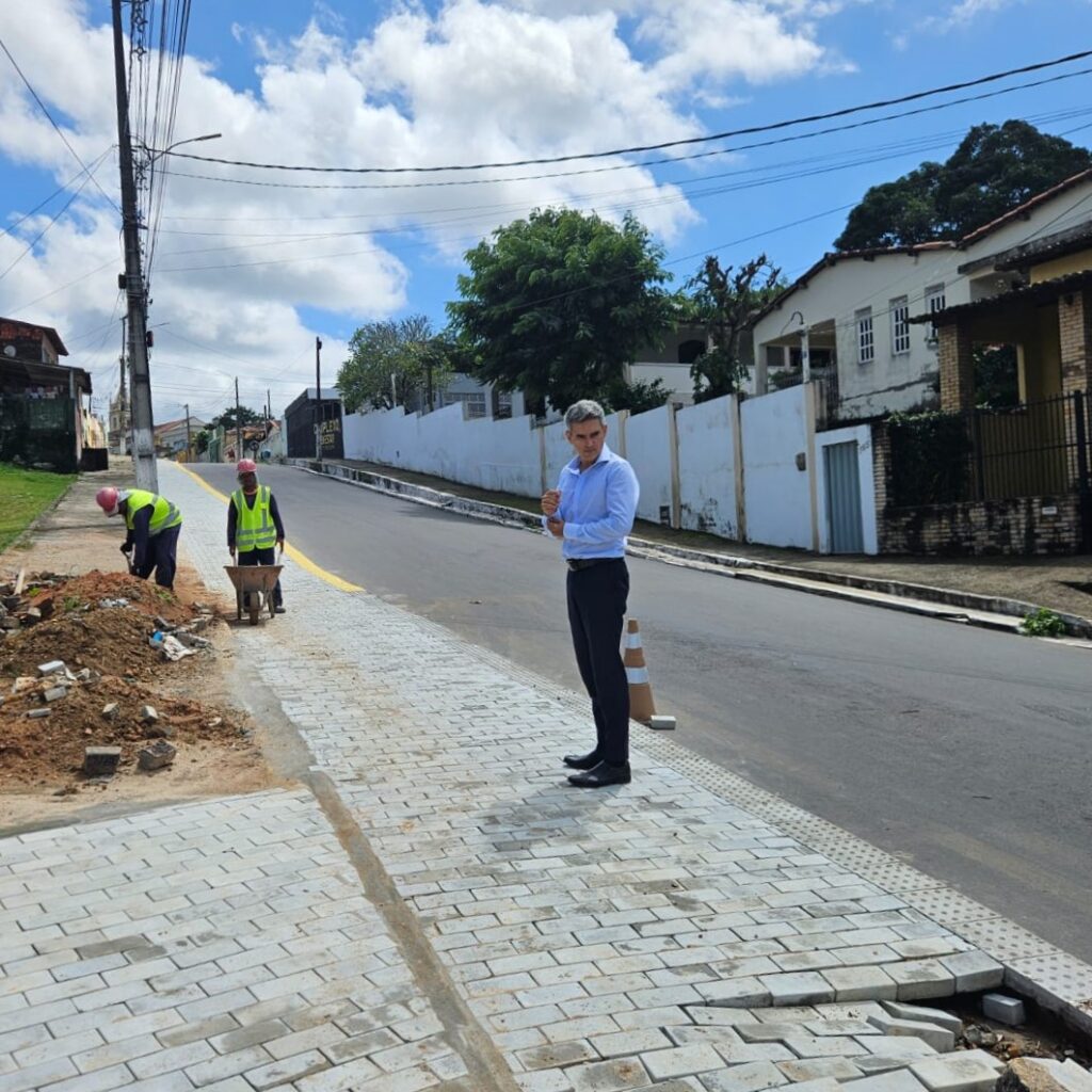 Homem de camisa social clara e calça escura observa dois operários trabalhando na pavimentação de uma calçada em uma rua residencial ladeada por casas e árvores sob céu ensolarado.