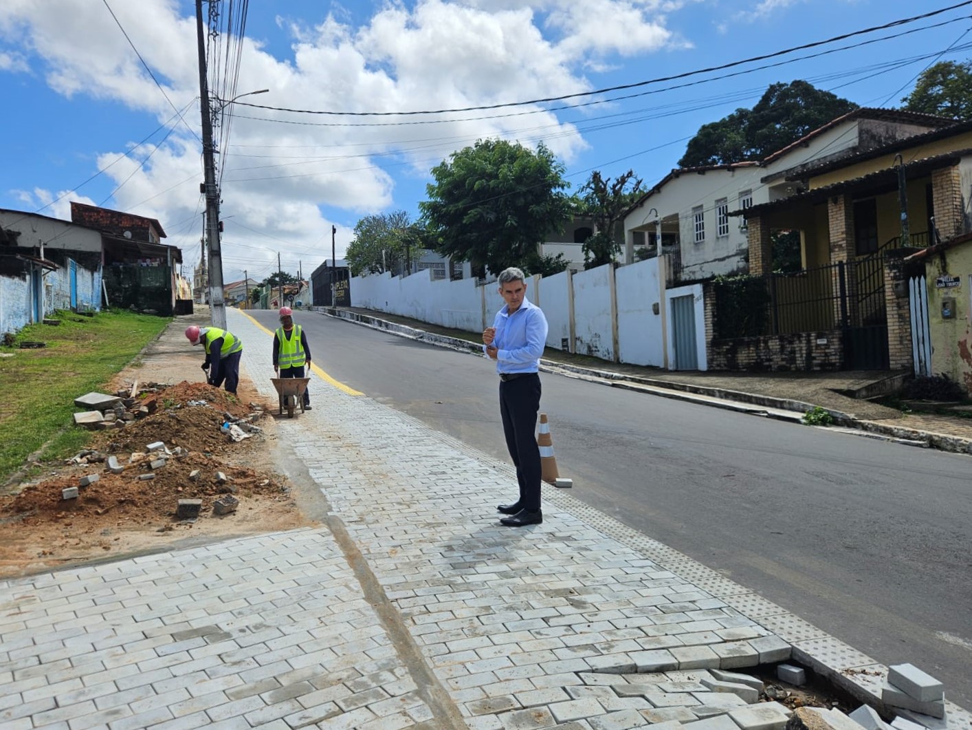 Homem de camisa social clara e calça escura observa dois operários trabalhando na pavimentação de uma calçada em uma rua residencial ladeada por casas e árvores sob céu ensolarado.