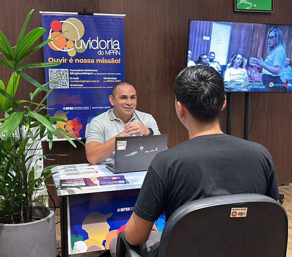 Homem sorridente atende um jovem sentado à sua frente em um guichê da Ouvidoria do Ministério Público do Rio Grande do Norte (MPRN), com um banner informativo e uma televisão ao fundo