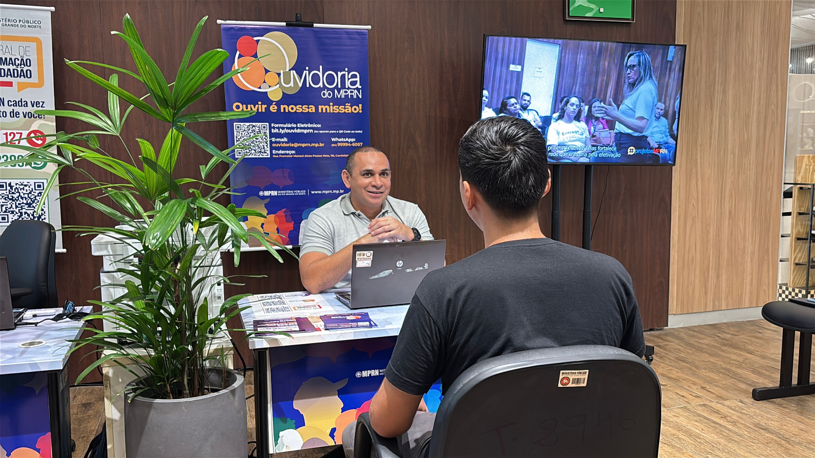 Homem sorridente atende um jovem sentado à sua frente em um guichê da Ouvidoria do Ministério Público do Rio Grande do Norte (MPRN), com um banner informativo e uma televisão ao fundo