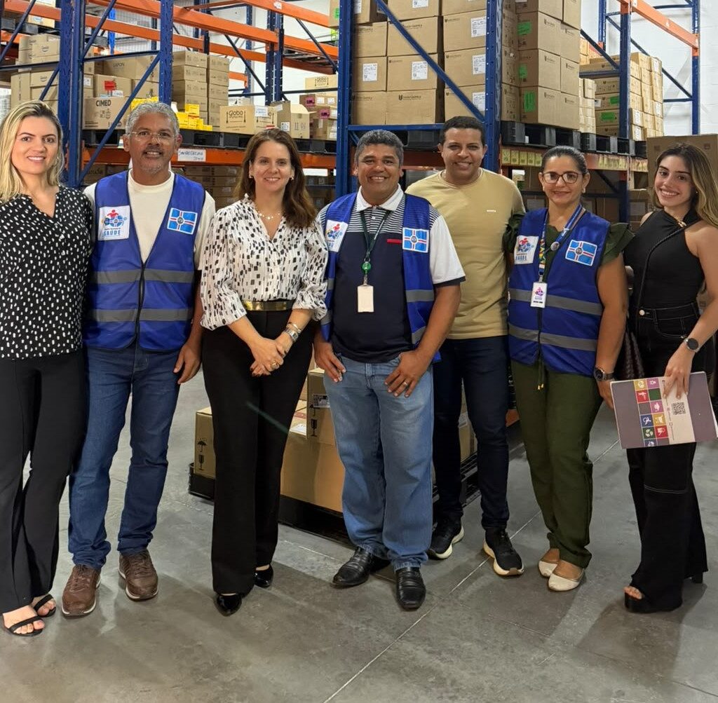 Grupo de sete pessoas sorri para a foto em pé lado a lado no interior de um galpão de armazenamento repleto de estantes com caixas.