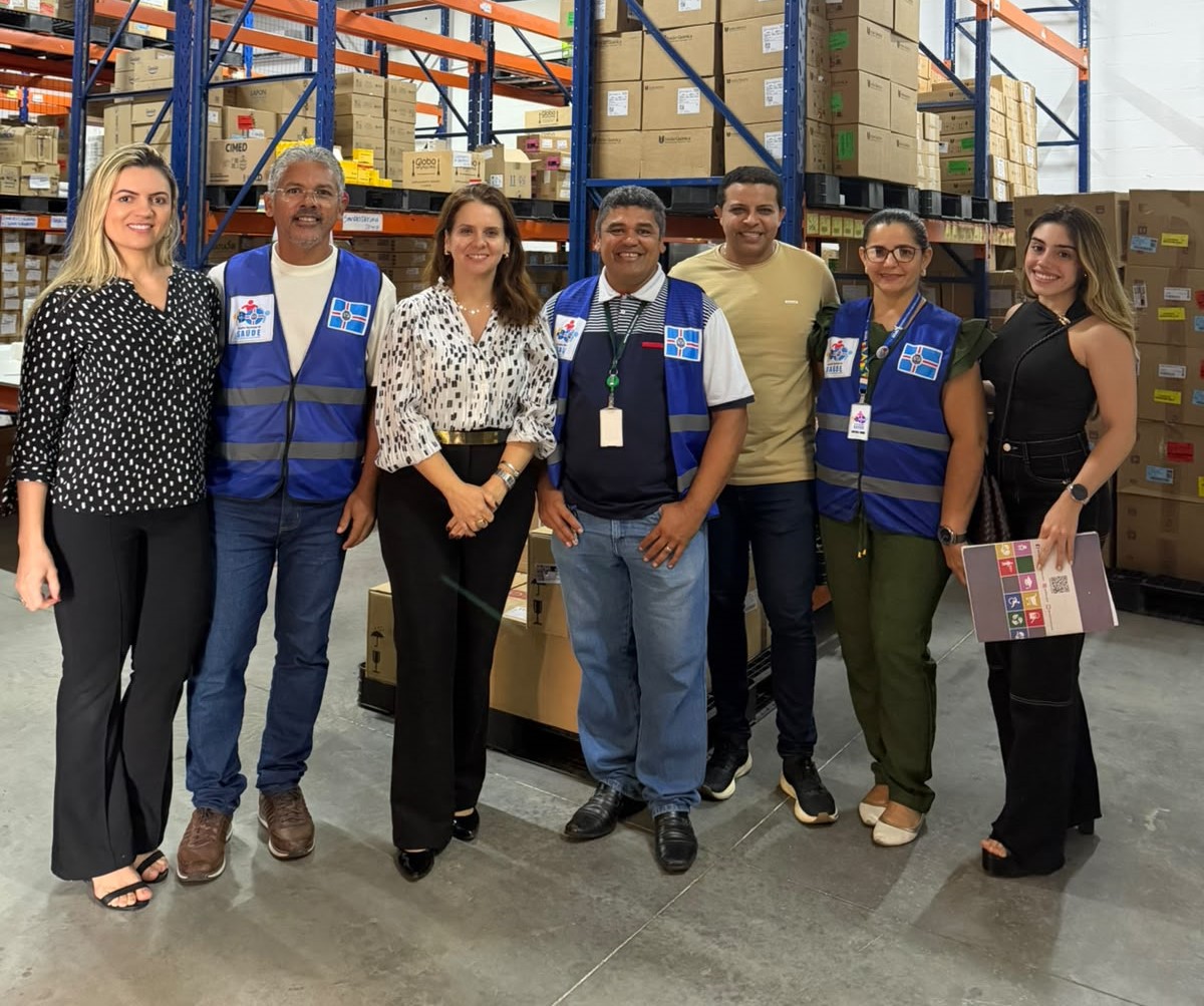 Grupo de sete pessoas sorri para a foto em pé lado a lado no interior de um galpão de armazenamento repleto de estantes com caixas.