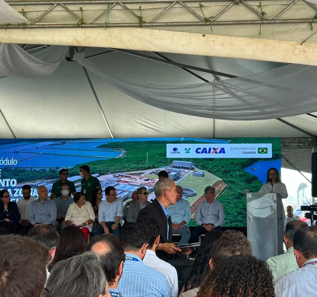 promotora discursa em um púlpito durante um evento oficial sob uma tenda, acompanhada por autoridades no palco e com uma grande plateia assistindo.