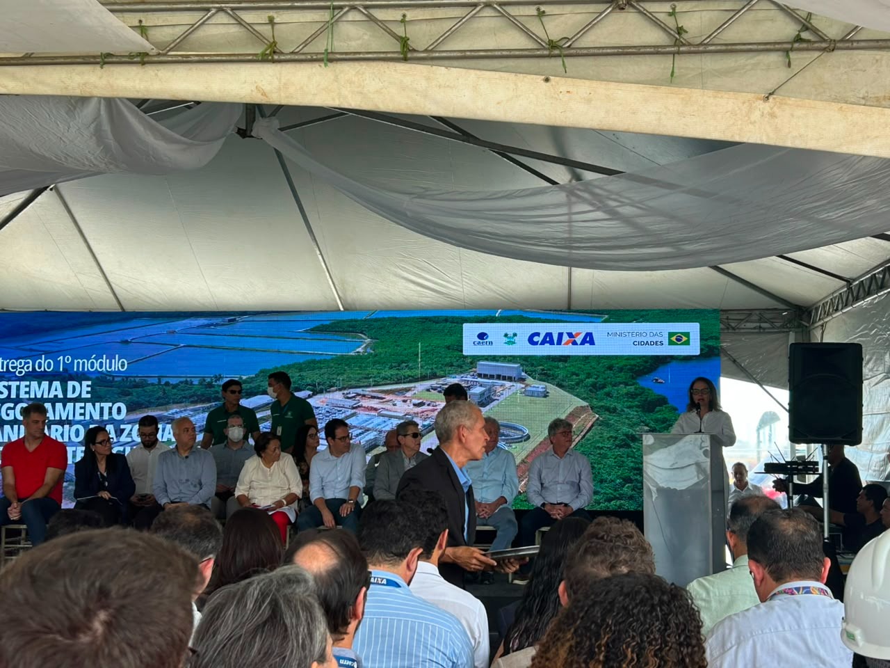 promotora discursa em um púlpito durante um evento oficial sob uma tenda, acompanhada por autoridades no palco e com uma grande plateia assistindo.