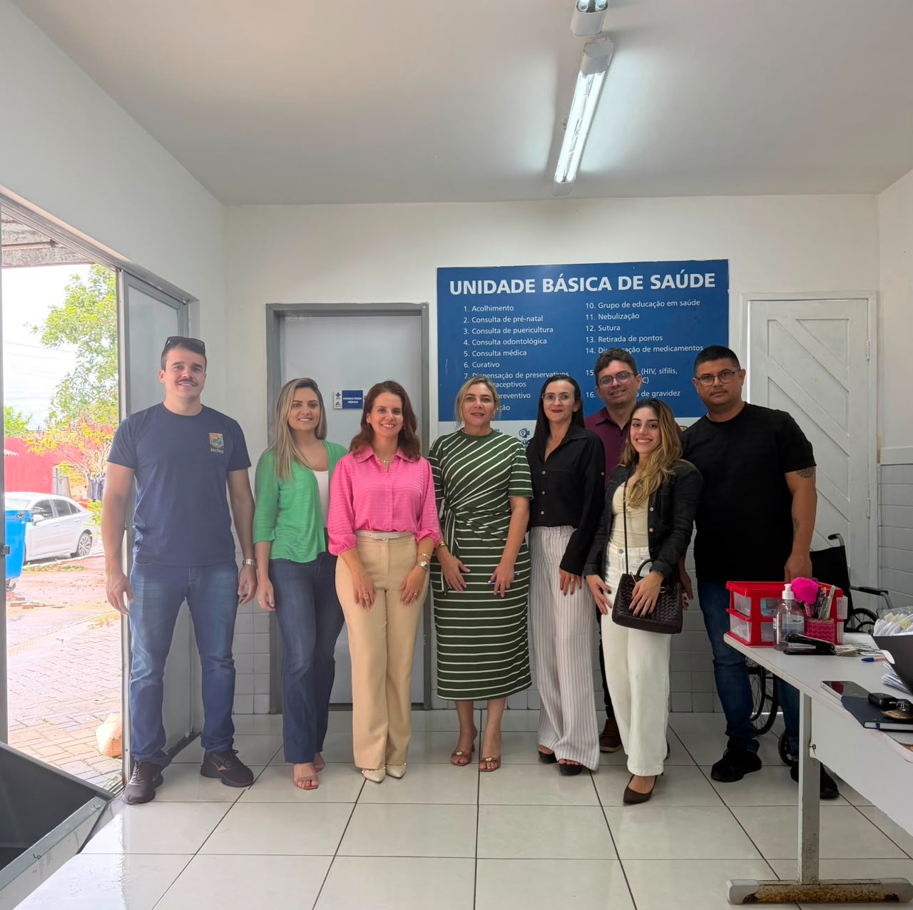 Um grupo de oito pessoas sorri em pé, lado a lado, dentro de uma Unidade Básica de Saúde, posando para uma foto em frente a uma placa azul com a lista de serviços oferecidos pelo local.