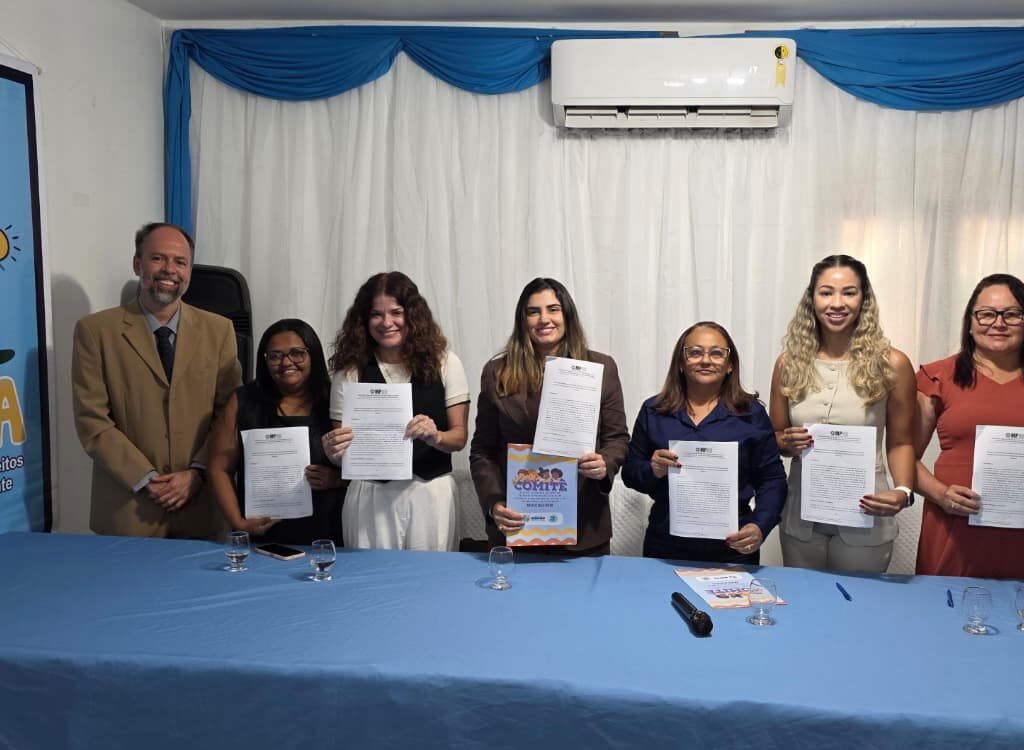 Sete pessoas estão lado a lado atrás de uma mesa azul, sorrindo e segurando documentos impressos em um evento oficial do CMDCA de Macau.