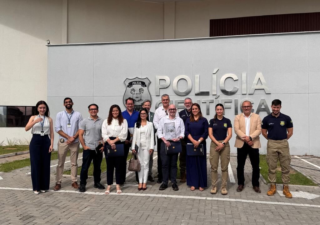 grupo de treze pessoas sorri e posa para foto em frente ao prédio da Polícia Científica.