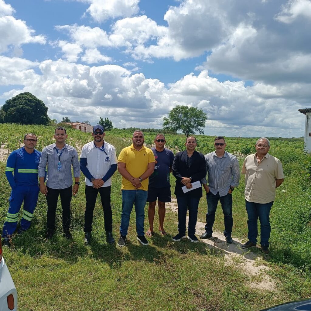 Grupo de oito homens em pé lado a lado em uma área rural ao ar livre, com vegetação e céu ensolarado ao fundo.
