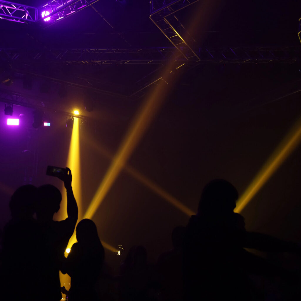 Silhuetas de pessoas em um show ou festa escura, iluminadas por feixes de luz coloridas
