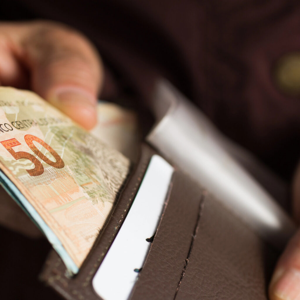 close mostra as mãos de uma pessoa idosa retirando uma nota de 50 reais de dentro de uma carteira marrom aberta