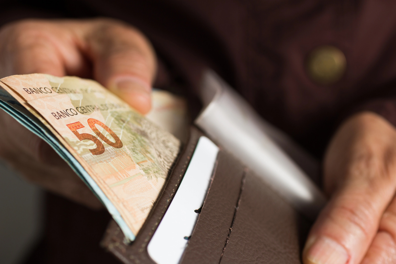 close mostra as mãos de uma pessoa idosa retirando uma nota de 50 reais de dentro de uma carteira marrom aberta