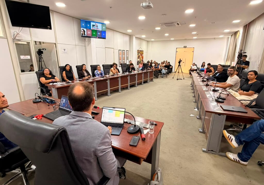 Pessoas reunidas em uma sala de conferências, dispostas ao redor de grandes mesas organizadas em formato de U, acompanhando uma reunião com transmissão ao vivo.