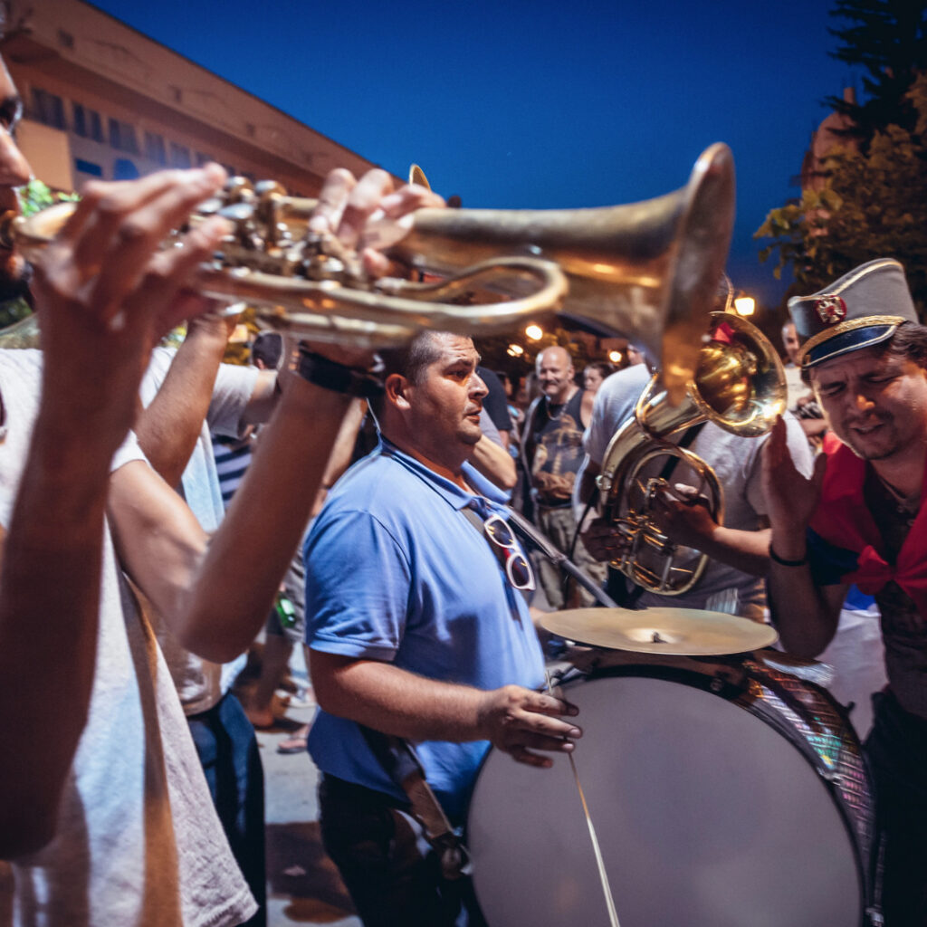 grupo de músicos toca trompete e percussão em uma festa ao ar livre à noite, cercado por pessoas.
