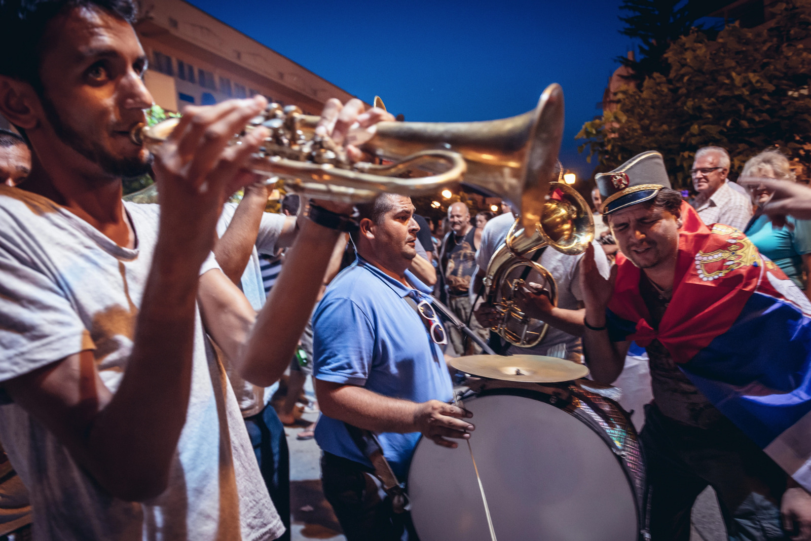 grupo de músicos toca trompete e percussão em uma festa ao ar livre à noite, cercado por pessoas.