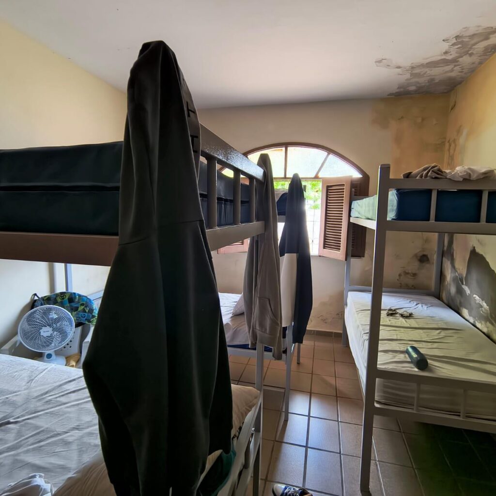 quarto compartilhado simples com várias beliches de metal, roupas penduradas nas camas, um ventilador de mesa no chão e uma janela arqueada ao fundo que ilumina o ambiente