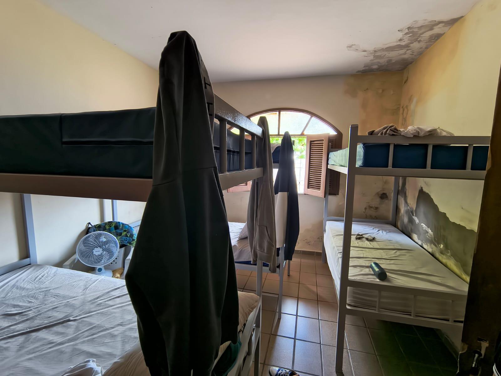 quarto compartilhado simples com várias beliches de metal, roupas penduradas nas camas, um ventilador de mesa no chão e uma janela arqueada ao fundo que ilumina o ambiente