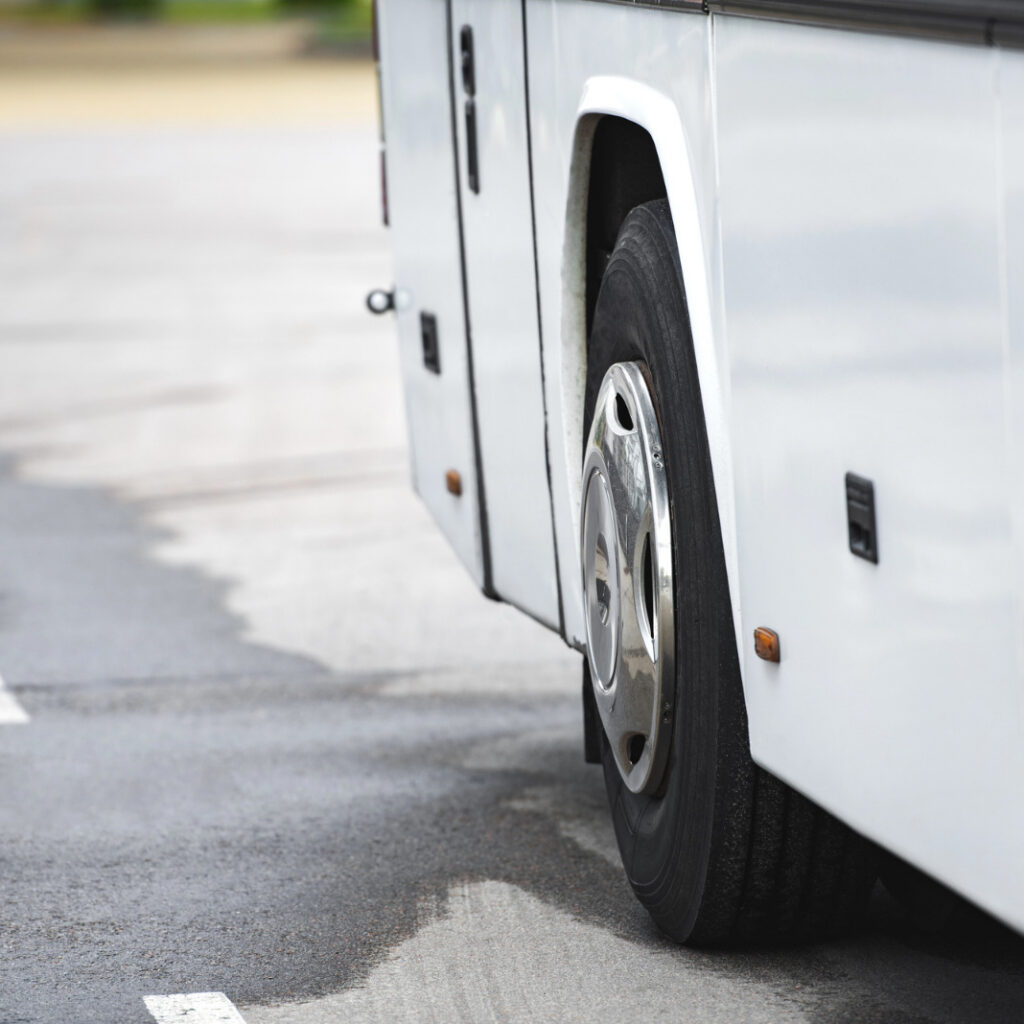 lateral inferior de um ônibus branco estacionado sobre o asfalto molhado, destacando um de seus pneus com calota cromada