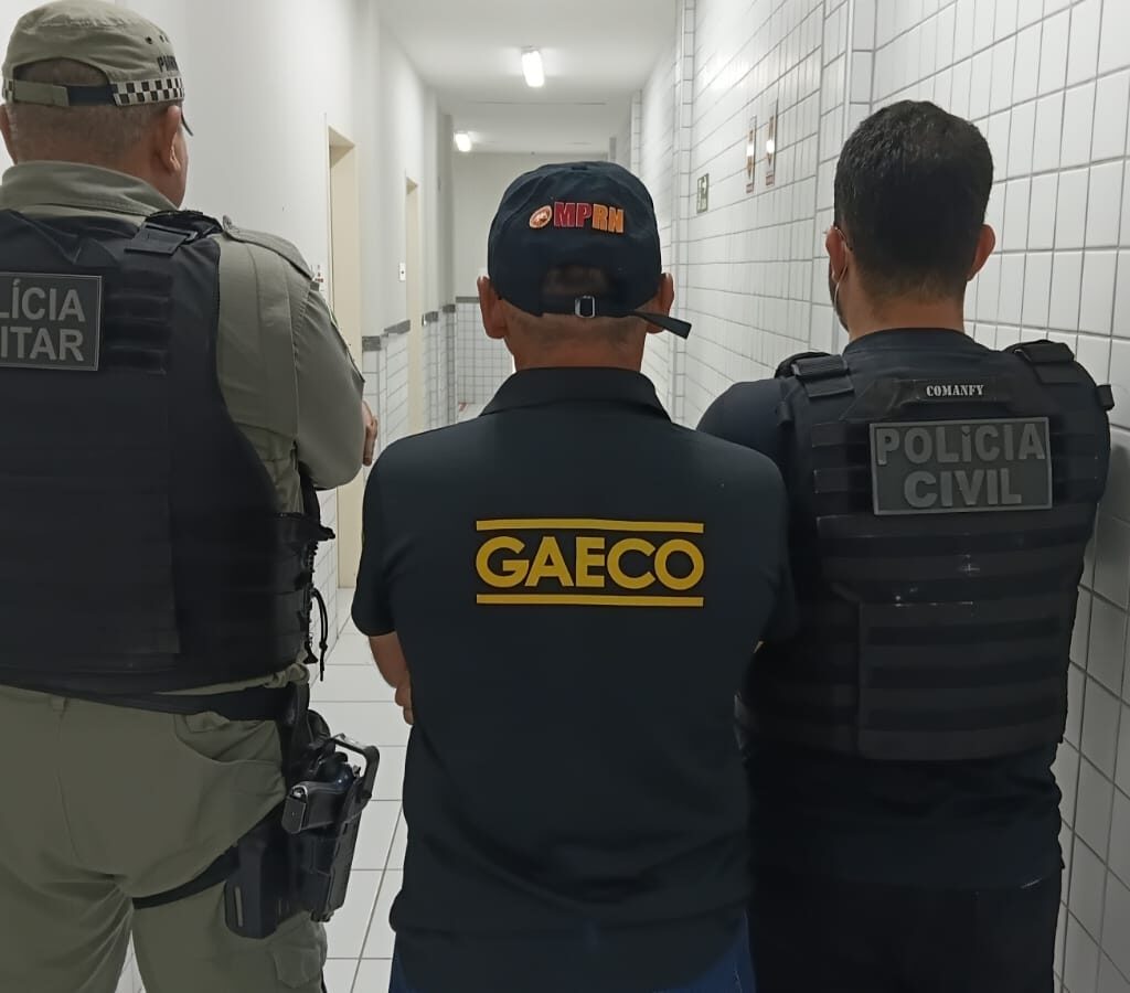 três homens de costas, sendo dois deles com farda da PM e um com uniforme e bone do gaeco/mprn