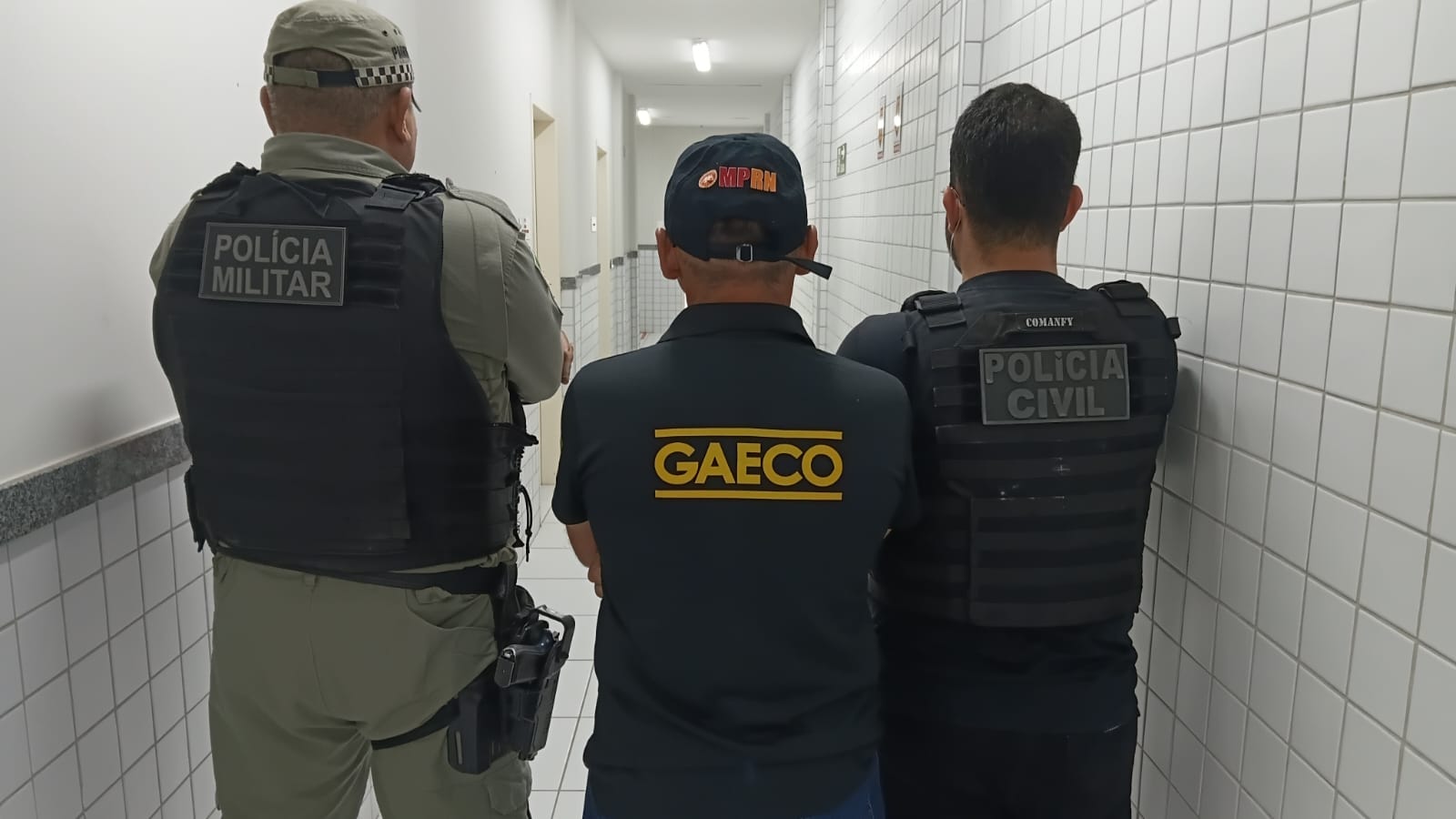 três homens de costas, sendo dois deles com farda da PM e um com uniforme e bone do gaeco/mprn