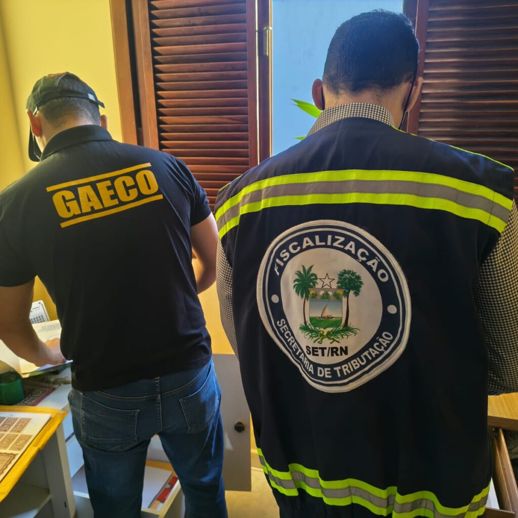 Dois agentes de fiscalização de costas, um usando uniforme do GAECO e o outro do SET/RN, examinam documentos em uma sala.