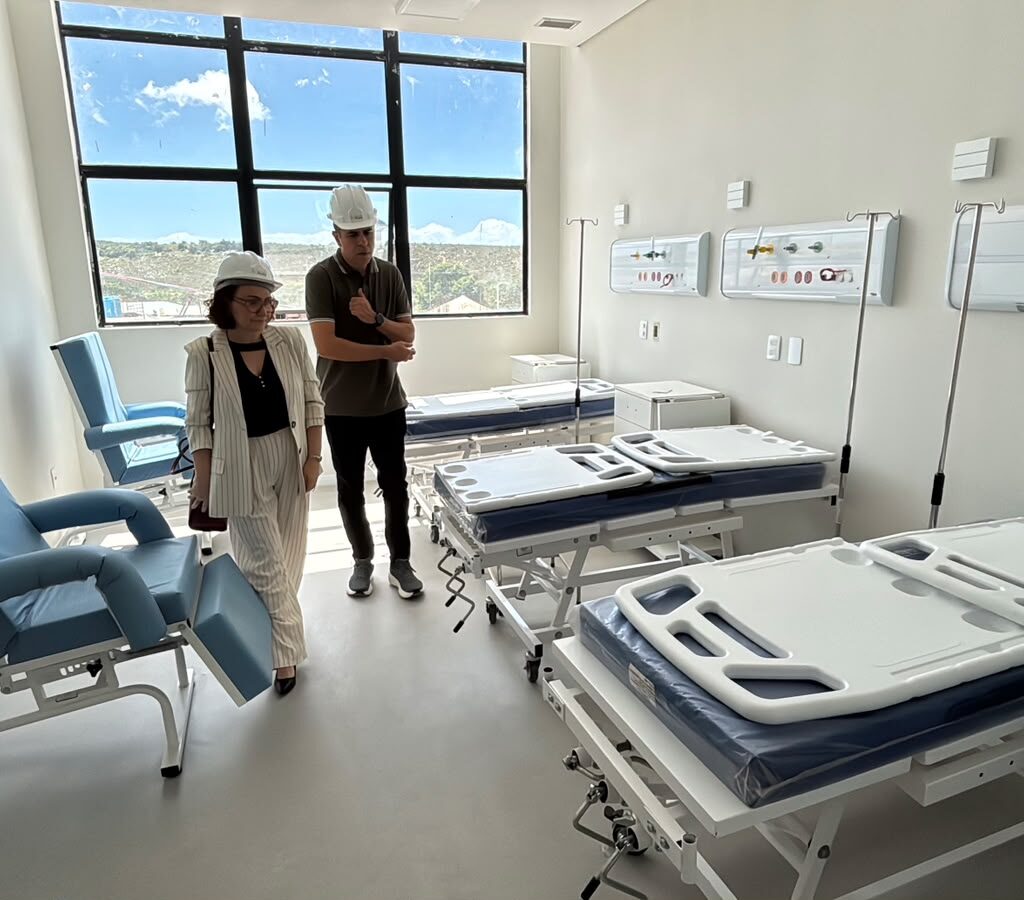 Duas pessoas usando capacetes brancos de proteção inspecionam um quarto hospitalar novo e iluminado, equipado com poltronas azuis e várias camas hospitalares dispostas em fila.