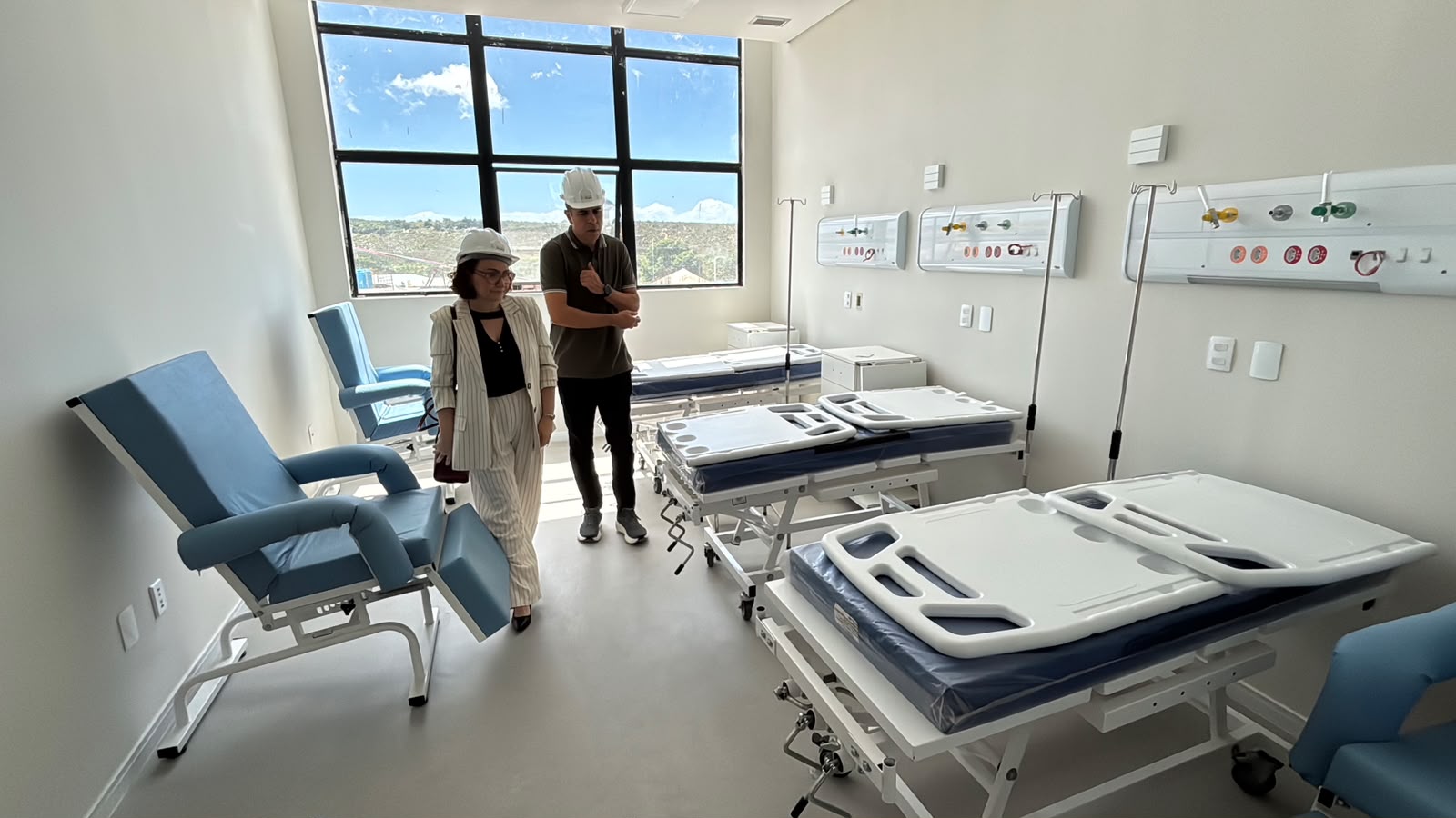 Duas pessoas usando capacetes brancos de proteção inspecionam um quarto hospitalar novo e iluminado, equipado com poltronas azuis e várias camas hospitalares dispostas em fila.