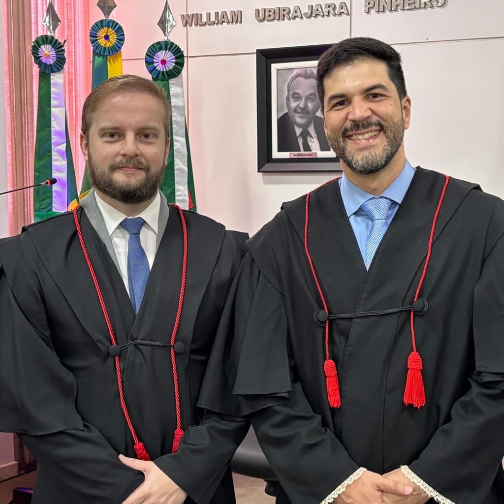 Dois homens vestidos com togas pretas estão em pé e posam para uma foto em um ambiente de escritório com bandeiras ao fundo.