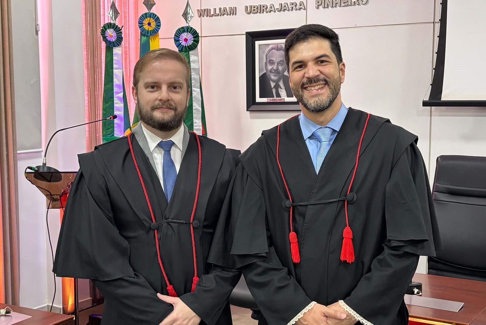 Dois homens vestidos com togas pretas estão em pé e posam para uma foto em um ambiente de escritório com bandeiras ao fundo.