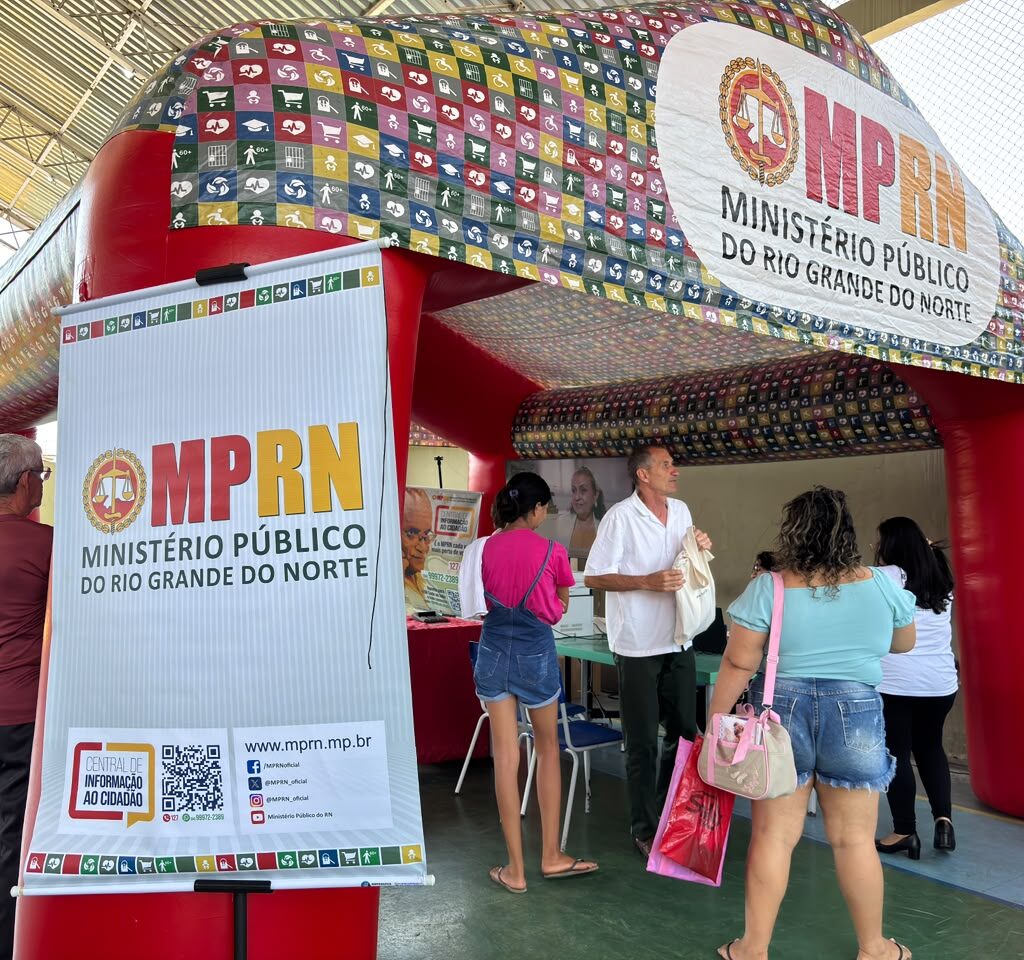 Uma tenda inflável vermelha do Ministério Público do Rio Grande do Norte (MPRN) está instalada em um espaço coberto, onde pessoas se aproximam do atendimento prestado por um homem com camisa branca.