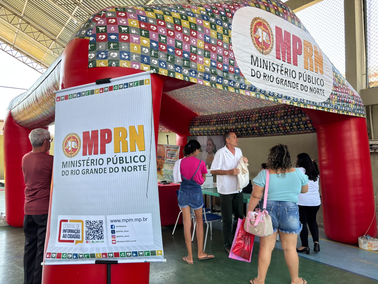 Uma tenda inflável vermelha do Ministério Público do Rio Grande do Norte (MPRN) está instalada em um espaço coberto, onde pessoas se aproximam do atendimento prestado por um homem com camisa branca.